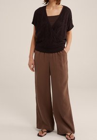 Donkerbruine wikkelstijl top met een gestructureerd patroon, gecombineerd met losse, lichtbruine wijde broek en zwarte teenflipflops.