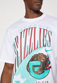 Λευκό T-shirt με γραφικό σχέδιο που περιλαμβάνει το κείμενο «Memphis Grizzlies», ένα λογότυπο αρκούδας και τόνους τιρκουάζ σε ανοιχτό μπλε φόντο.