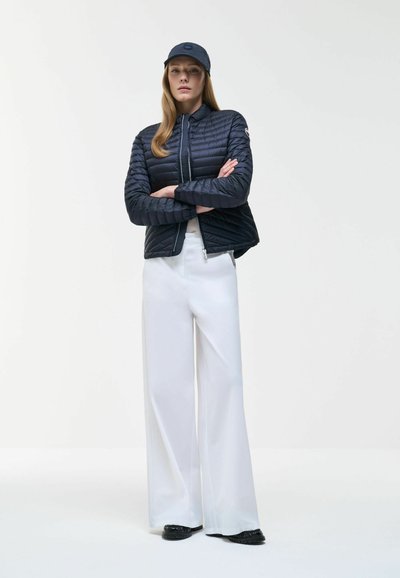 Woman wearing a black quilted jacket, matching cap, white wide-leg pants, and black shoes, standing with arms crossed on a plain background.