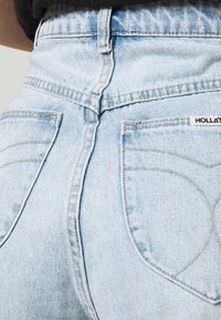 Jeans de ganga de cintura alta em azul claro com uma textura desgastada, apresentando dois bolsos traseiros com costuras curvas e um pequeno rótulo da marca.