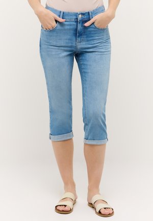 Vrouw die lichtblauwe opgerolde denim capri broek draagt met handen in de zakken en beige platte sandalen, staand op een witte achtergrond.