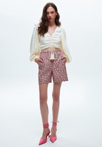 Weißes Blusenshirt mit elastischen Details und langen Puffärmeln, kombiniert mit rosafarbenen gemusterten Shorts und rosafarbenen Stiletto-Absätzen mit Wickelriemen.