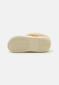 Chausson beige avec un dessus en textile doux et une semelle intérieure rembourrée. La semelle extérieure en caoutchouc blanc présente un motif de semelle antidérapant pour une meilleure adhérence.