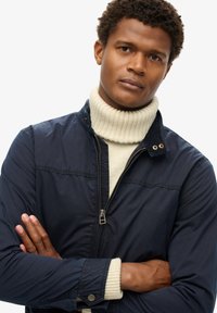 Marineblå jakke med lynlås, stående krave og knaplukning ved håndleddene, lagdelt over en cremefarvet turtleneck sweater. Glat tekstur, casual design.