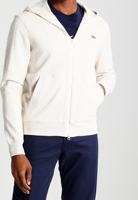 Sudadera con cremallera color crema, de tela texturizada, que cuenta con una cremallera frontal, dos bolsillos laterales y un pequeño logo verde en el lado izquierdo del pecho.