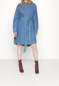 Denim shirt dress with long sleeves, button closure, and a belted waist. Features a rounded hem and contrast stitching. Worn with burgundy ankle boots.