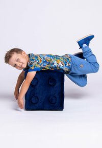 Niebieski pluszowy sześcian z okrągłymi akcentami; dziecko w kolorowej koszuli w wzory leży na górze, w niebieskich spodniach i trampkach.