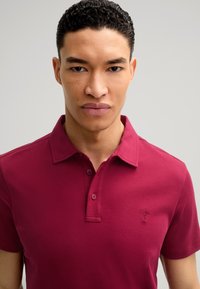 Burgunderfarbendes Polo-Shirt mit kurzen Ärmeln aus glattem Baumwollmaterial, mit Kragen, Knopfleiste und kleinem gesticktem Logo auf der Brust.