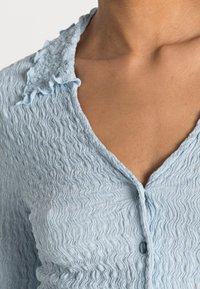 Gros plan sur une personne portant un haut texturé bleu clair avec un col volanté et un seul bouton visible au niveau de l'encolure.