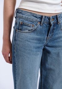 Jean en denim bleu clair avec une taille haute, une coupe droite et des coutures visibles. Comprend des poches avant avec des rivets en cuivre.