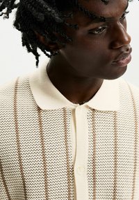 Knit polo shirt in cream with a textured pattern and brown stripes. Features a ribbed collar and buttoned placket. Soft and breathable material.