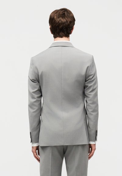 Άνδρας που φοράει ανοιχτό γκρι κοστούμι με λευκό πουκάμισο ορατό στους μανσέτες, στέκεται γυρισμένος πλάτη σε ένα απλό ανοιχτό φόντο.
