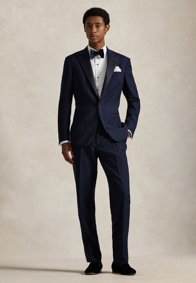 Man wearing navy blue tuxedo with black bow tie, white dress shirt, and black dress shoes, standing against plain beige background.
