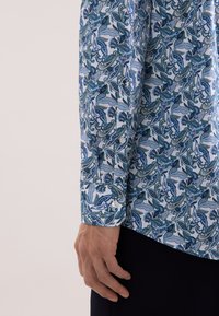 Lang shirt met lange mouwen, voorzien van een blauw-groen paisley-patroon op een lichte achtergrond. Het materiaal lijkt glad en heeft knoopmanchet details.