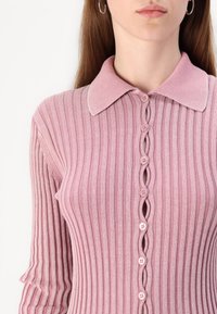 Sweater rosa canelado com gola, apresentando um fecho de botão ondulado e mangas longas, feito de um material suave e texturizado.