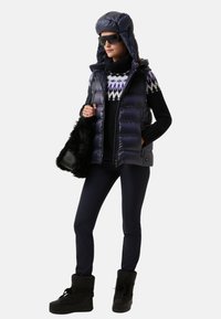 Frau trägt eine navy farbene gepolsterte Weste und passenden Hut, einen schwarz gemusterten Pullover, dunkle Hosen, schwarze Stiefel, eine Sonnenbrille und eine schwarze Pelztasche.