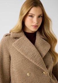 Beiger zweireihiger Mantel aus strukturiertem Stoff mit weitem Kragen, versehen mit goldenen Knöpfen und einer weichen, flauschigen Textur.