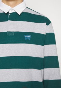 Groen en grijs gestreepte longsleeve polo shirt, met een witte kraag en een blauwe Wrangler logo patch op de borst. Zachte stof textuur.