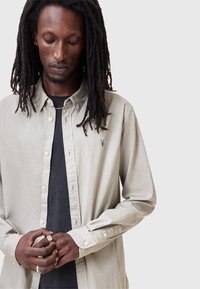 Lichtbeige button-up shirt met een klassieke kraag, lange mouwen en een subtiele logodetail. Draagt over een zwart t-shirt. Gladde textuur.