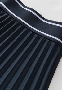 Jupe plissée marine avec une taille solide ornée d'une bande blanche. Le tissu présente une texture lisse et un plissé structuré.