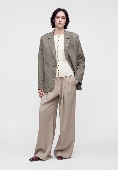 Femme portant un blazer à carreaux, un cardigan crème boutonné, un pantalon large beige et des chaussures pointues bordeaux, debout avec une main dans la poche.
