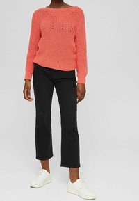 Koraliowy sweter dzianinowy z szerokim dekoltem w łódkę i dekoracyjnym wzorem ażurowym, zestawiony z czarnymi dżinsami i białymi trampkami.