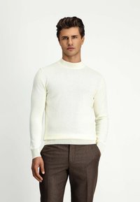 Cremfarbener Strickpullover mit langen Ärmeln und hohem Kragen. Das Material wirkt glatt und ist kombiniert mit braunen, strukturierten Hosen.