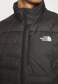 Giacca nera isolata con colletto alto, zip frontale e texture trapuntata. Presenta un piccolo logo bianco sul lato sinistro del petto.