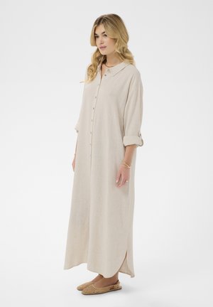 Žena s dlhými blond vlasmi nosí béžové maxi šaty na gombíky s dlhými rukávmi a vyhrnutými manžetami, a béžové pletené slingback baleríny, stojaca zboku.