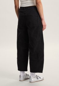 Pantalones anchos de mezclilla negra con cintura alta, detalles de costuras visibles y dos bolsillos traseros, combinados con zapatos plateados y calcetines blancos.