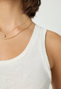 Nahaufnahme einer Person, die ein weißes ärmelloses Oberteil und zwei Goldketten trägt, eine davon mit einem kleinen runden Anhänger.