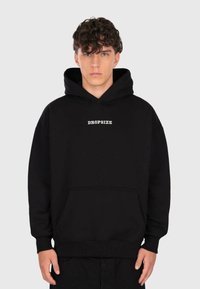 Junger Mann mit lockigem Haar, der einen schwarzen Hoodie mit dem Text "DROPSIZE" auf der Brust trägt, steht vor einem einfarbigen hellen Hintergrund.