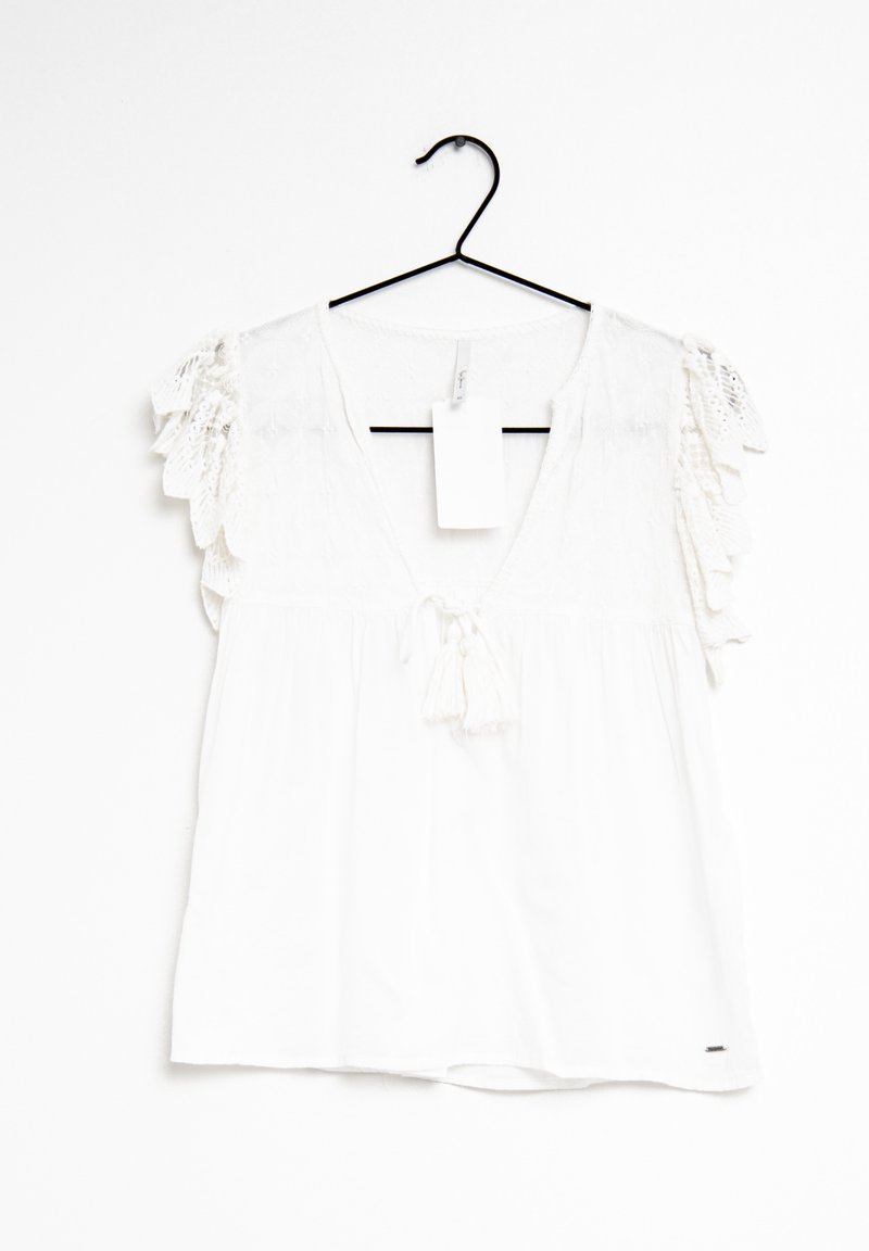 Blouse blanche avec manches courtes en dentelle, col en V profond, lien à pompons, et une étiquette blanche vierge suspendue à un cintre noir sur un fond blanc.