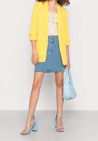 Žlutý blejzr s ohrnutými rukávy přes květinové tričko, v kombinaci s džínovou sukní a světle modrými doplňky, včetně podpatků a kabelky.