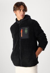 Giacca in pile nera con zip frontale, cappuccio e tasca sul petto. Presenta una zip arancione e un design grafico verde sulla tasca. Superficie testurizzata.