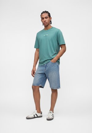 Junger Mann mit geflochtenem Haar, der ein türkisfarbenes Grafik-T-Shirt, hellblaue Denim-Shorts und weiße Sneakers mit schwarzen Streifen trägt, steht mit einer Hand in der Hosentasche.