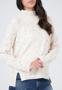 Off-white gebreide trui met een hoge ribbelkraag, gesprenkeld zwarte accenten, zijsplitten en ribbelzoom, gecombineerd met blauwe jeans.