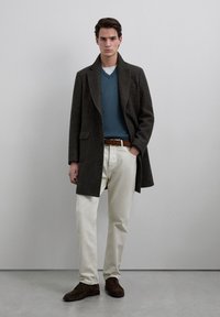 Bruine wollen jas over een blauwe V-halstrui, gecombineerd met lichtbeige broek en bruine suède schoenen. Eenvoudig ontwerp met zakdetail.