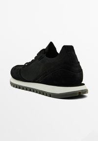 Scarpa sportiva nera realizzata con una combinazione di rete e suede. Presenta una parte superiore testurizzata, un collo imbottito e una suola in gomma bianca con scanalature.