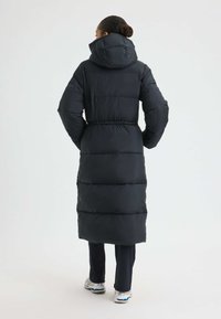 Schwarzer, gepolsterter langer Mantel mit Kapuze, ausgestattet mit einem gesteppten Design und einem elastischen Bund. Kombiniert mit dunklen Hosen und mehrfarbigen Sneakers.