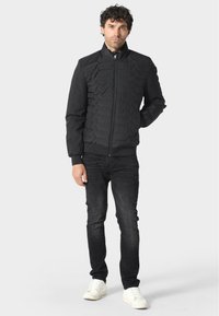 Czarna kurtka puchowa z fakturowanym pikowanym wzorem, wysoka szyja z zamkiem, noszona na ciemnych dżinsach i białych trampkach. Gładkie wykończenie materiału.