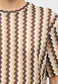 Πλεκτό τοπ με ζιγκ ζαγκ σε αποχρώσεις ανοιχτού μπεζ, καφέ και λευκού, με κοντά φουσκωτά μανίκια και στρογγυλή λαιμόκοψη.
