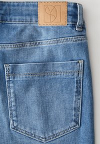 Jeans in denim azzurro chiaro con una toppa in pelle color tan caratterizzata da un design in rilievo, tasca posteriore con dettagli di cucitura sottile. Texture liscia.