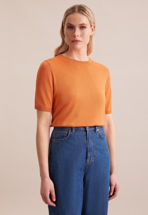 Vrouw met blond haar die een oranje korte mouwen trui draagt, ingestopt in een hooggetailleerde blauwe spijkerbroek, tegen een effen beige achtergrond.