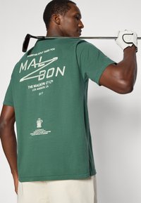 Πράσινο βαμβακερό μπλουζάκι με λευκό κείμενο και γραφικά στην πλάτη, κοντά μανίκια και άνετη εφαρμογή. Ένα γκολφ κλαμπ ακουμπάει στον ώμο.