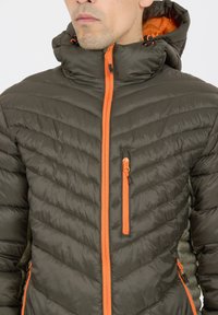 Oliven grøn pufferjakke med en quiltet design, lys orange lynlås og detaljer, justerbar hætte, og to lynlåslommer foran.