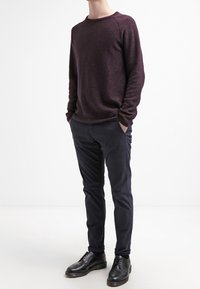 Muž v tmavě vínovém svetru, černých slim-fit kalhotách a černých formálních botách, stojící s rukama v kapsách na bílém pozadí.