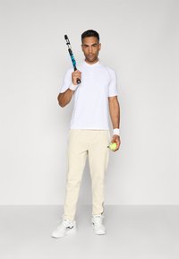Chemise polo blanche à manches courtes avec détails de boutons, associée à un pantalon beige. Le sujet tient une raquette de tennis et une balle de tennis verte. Baskets blanches.