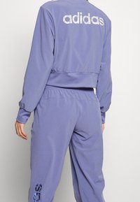 Lila idrottsjacka och byxor i ett set av mjukt tyg, med en kortad design och "adidas"-logotypen i reflektivt material på baksidan.