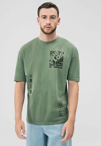 Olivgrünes Baumwoll-T-Shirt mit Blockdruck-Grafiken in Schwarz, rundem Ausschnitt, kurzen Ärmeln und Textdetails auf der Vorderseite und dem Ärmel.
